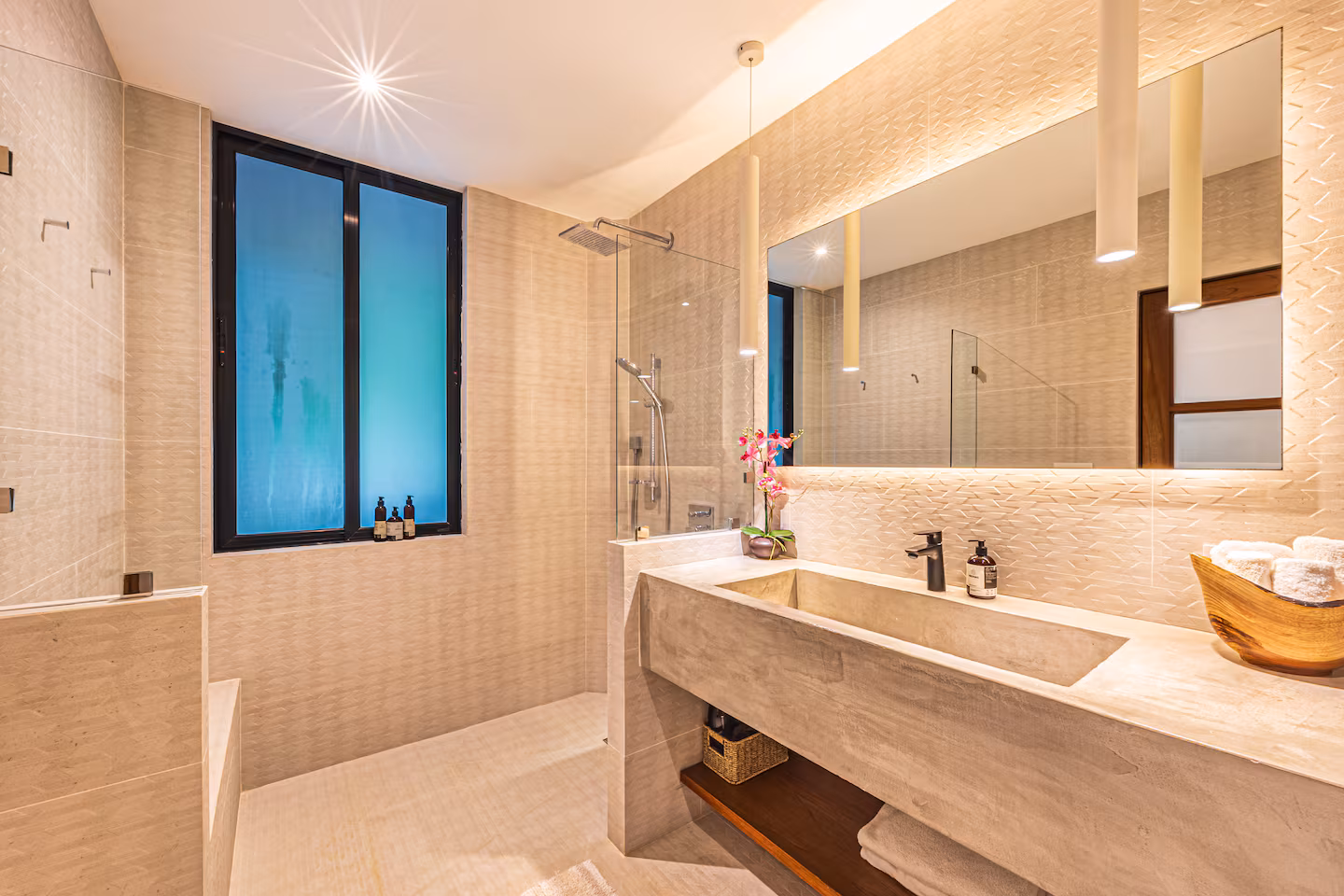 Spa-style bathroom with concrete vanity, glass shower, textured tile walls, and soft pendant lighting in a villa near Dominical, Costa Rica.