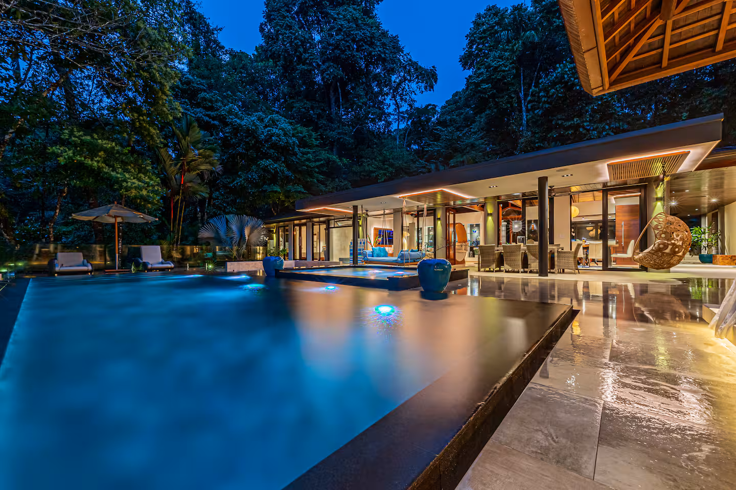 Infinity pool at night with soft lighting, open living areas, and dense jungle surrounding the villa.