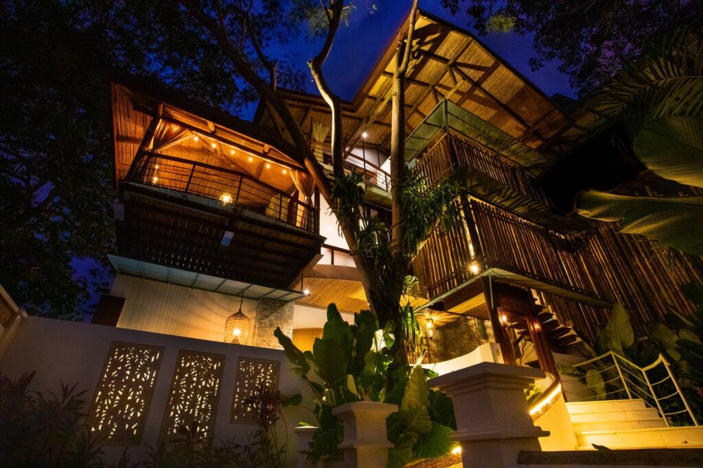 As night falls, warm lighting reveals the villa’s layered architecture, creating an inviting glow amid the surrounding tropical landscape.