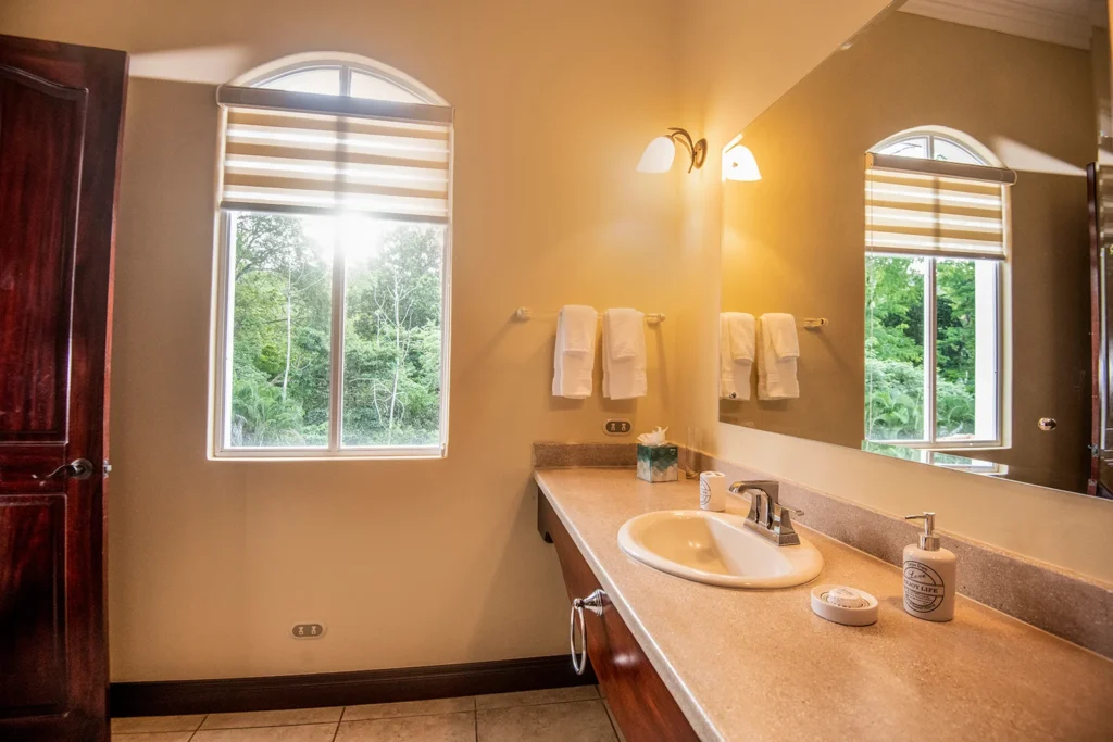 A bright bathroom vanity with a large mirror and a window overlooking tropical greenery.