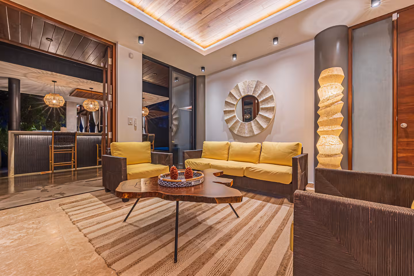 Indoor lounge with yellow seating, wood ceiling, decorative lighting, and open access to a bar area.