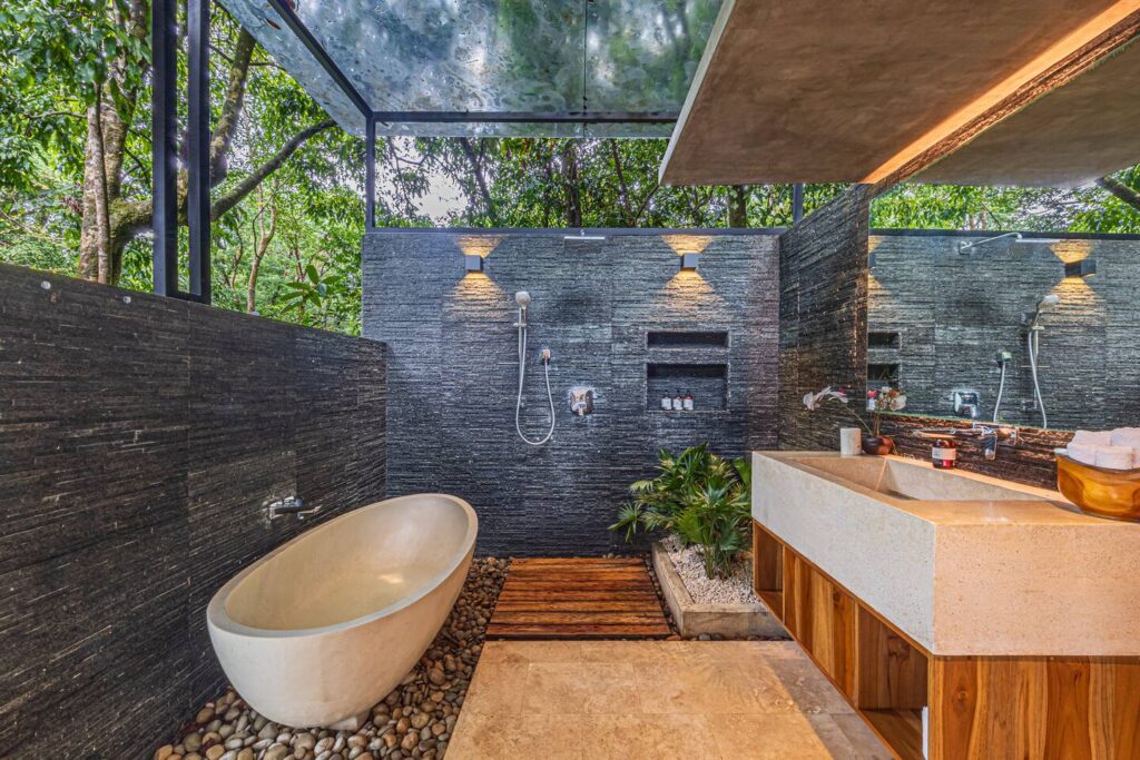 An open-air spa bathroom framed by large windows, blending stone textures with immersive jungle views.