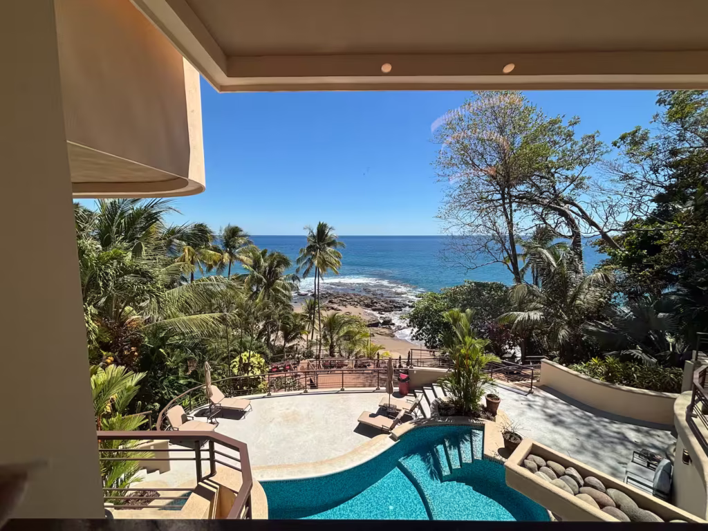The pool terrace overlooks the Pacific Ocean, with tropical gardens, a beachfront deck, and the shoreline just beyond.