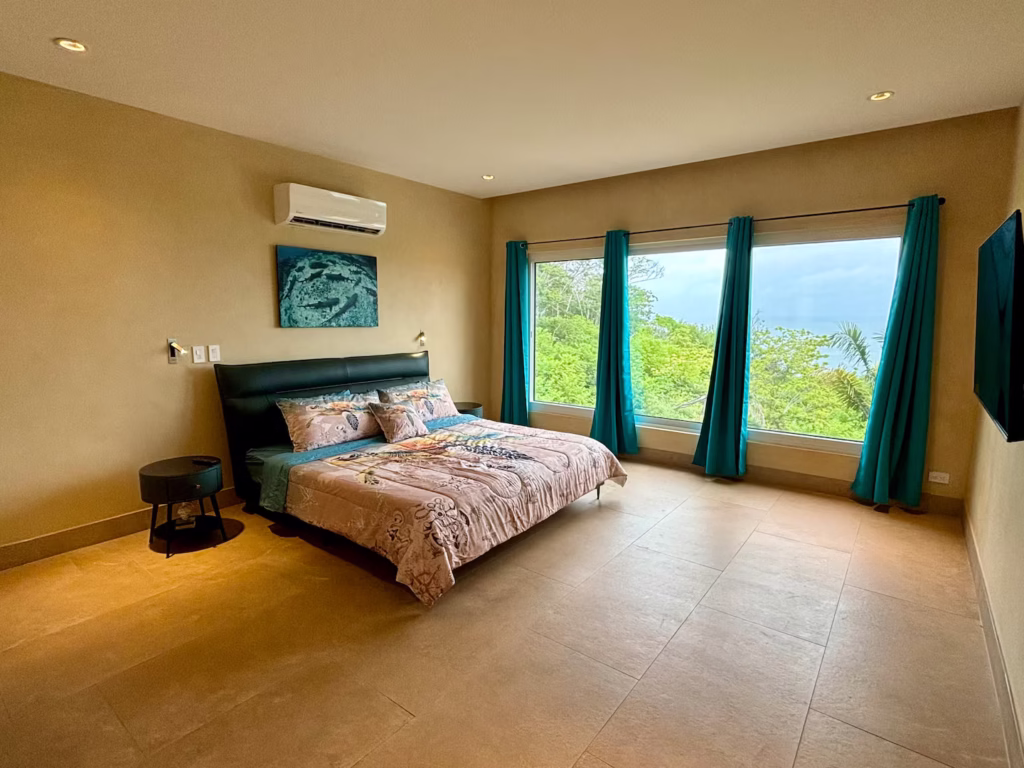 A spacious king bedroom features large windows overlooking the tropical forest with partial Pacific Ocean views.
